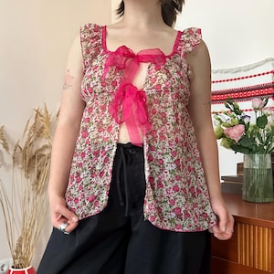 Pode incluir: Uma blusa sem mangas transparente com um padrão floral vibrante em rosa e verde sobre um fundo branco. A peça possui um decote quadrado, detalhes de folhos nos ombros e uma abertura frontal presa por dois laços de fita rosa translúcida.