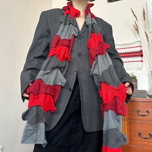 Puede incluir: Una bufanda larga y en capas en tonos rojos y grises. La bufanda está hecha de un material suave y texturizado y tiene un diseño con volantes. La bufanda se lleva sobre una blazer gris oscuro.