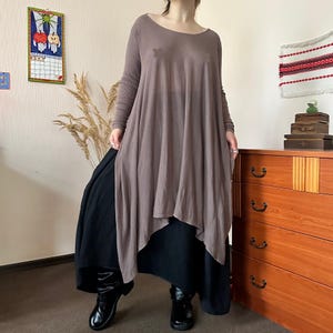 May include: A long-sleeved, taupe-colored tunic with a scoop neckline and an asymmetrical hemline, layered over a solid black skirt. The outfit is paired with black boots, creating a flowing silhouette with visible fabric texture.