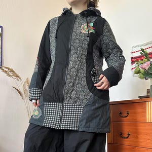 Könnte beinhalten: Eine Kapuzenjacke mit Reißverschluss, die aus schwarzen Stoffbahnen, grauem Damastmuster und schwarz-weiß karierten Vichy-Abschnitten besteht. Die Jacke ist mit bunten, kreisförmigen Stickereien auf der Brust und unten links versehen.