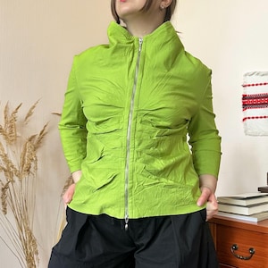 May include: A bright green jacket with a high collar and a full-length silver zipper. The long-sleeved jacket has a ruched design and is paired with black pants. The jacket is a vibrant, eye-catching piece.