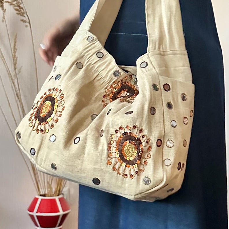 May include: A beige hobo-style shoulder bag crafted from textured fabric, featuring circular embroidered floral motifs in shades of brown, orange, and gold sequins. The exterior is decorated with small, reflective circular mirrors.