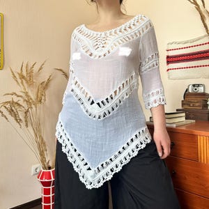 May include: White crochet top with a V-neck design and three-quarter sleeves. The top features intricate crochet detailing along the neckline, sleeves, and hem. The top is paired with black pants.