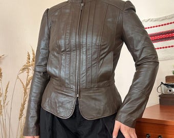 May include: A dark brown leather jacket featuring a high mandarin collar, a full-length front zipper closure, and vertical stitched paneling details on the chest. The jacket has long sleeves and a slightly flared peplum hemline at the waist.