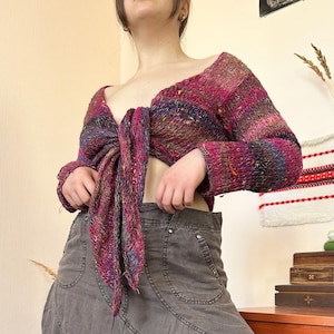 May include: A multi-colored, long-sleeved knit top with a tie closure. The top features shades of pink, purple, and blue, with a textured appearance. The sleeves are long, and the top is designed to be tied at the waist. The garment is a unique piece of clothing.