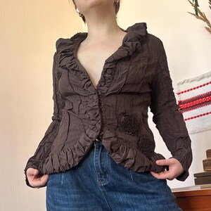 Può includere: Giacca marrone strutturata con finiture a balze e abbellimenti floreali. La giacca ha maniche lunghe e chiusura con bottoni. Lo stile è vintage e unico.