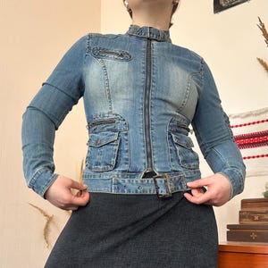 May include: A light blue denim jacket with a stand-up collar, zip fastening, and multiple pockets. The jacket has a belt at the waist and a fitted shape. The sleeves are long and the denim has a faded wash.