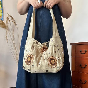 May include: A beige hobo-style shoulder bag made of fabric, featuring circular floral-like embroidery with brown and gold sequins. The bag is decorated with small, reflective circular mirrors scattered across the surface and has a wide shoulder strap.