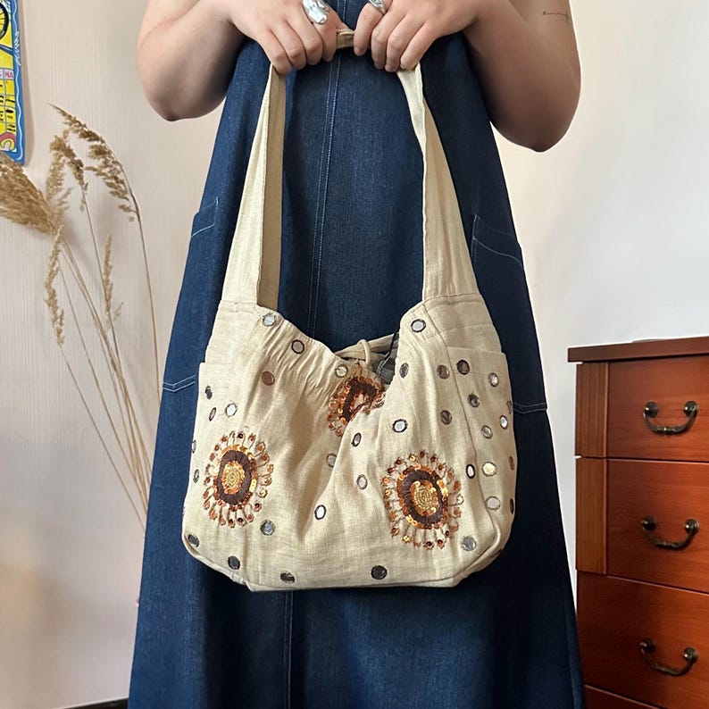 May include: A beige hobo-style shoulder bag made of fabric, featuring circular floral-like embroidery with brown and gold sequins. The bag is decorated with small, reflective circular mirrors scattered across the surface and has a wide shoulder strap.