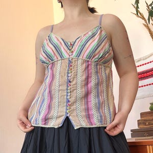 Könnte beinhalten: Ein bunt gestreiftes Camisole-Top mit V-Ausschnitt und dünnen Trägern. Das Top hat vertikale Streifen in verschiedenen Farben, darunter Rosa, Blau und Beige, und ist mit kleinen, bunten Knöpfen in der Mitte verziert. Der untere Teil ist einfarbig dunkel.