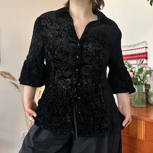 May include: A black button-down shirt featuring a tonal velvet paisley pattern, a collared neckline, and three-quarter length sleeves with flared cuffs. The garment is constructed with a textured fabric and a central vertical row of small buttons.