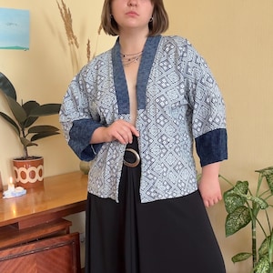 May include: A light blue and white patterned kimono-style jacket with dark blue trim on the collar and sleeves. The jacket is open in the front and has a geometric design. The person is wearing a black skirt.