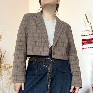 Puede incluir: Chaqueta corta a cuadros marrones con un solo botón. La chaqueta tiene una solapa con muescas clásica y se combina con una falda vaquera y un cinturón marrón. Un camiseta gris completa el conjunto.