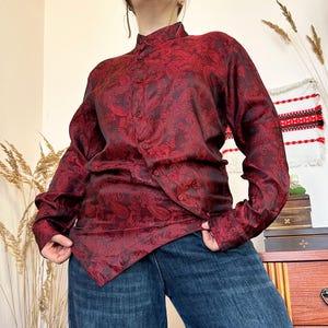 May include: A burgundy paisley print button-up shirt with long sleeves. The shirt has a mandarin collar and a unique asymmetrical button closure. The fabric has a subtle sheen, and the paisley pattern is a darker shade of burgundy.