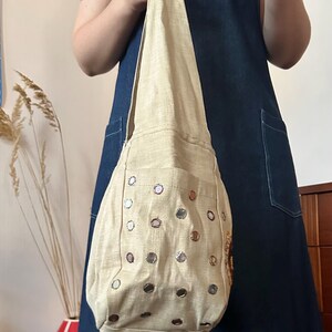 May include: A beige hobo-style shoulder bag made of a textured fabric, featuring a single integrated strap and a front pocket adorned with numerous small, circular reflective mirrors arranged in a grid pattern across the surface of the bag.