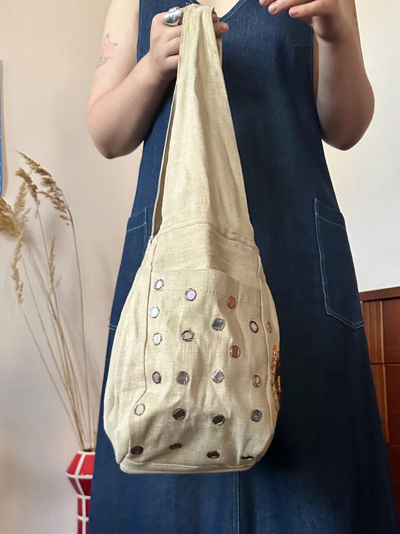 May include: A beige hobo-style shoulder bag made of a textured fabric, featuring a single integrated strap and a front pocket adorned with numerous small, circular reflective mirrors arranged in a grid pattern across the surface of the bag.