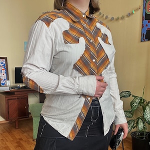 Può includere: Una camicia a maniche lunghe con un design patchwork unico. La camicia presenta una base beige con accenti a quadri arancioni, marroni e grigi. Le maniche e il corpo hanno un aspetto strutturato e la camicia è abbottonata.