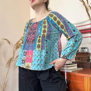 Op de afbeelding: Een patchwork blouse met een vierkante halslijn en lange mouwen. De blouse heeft een mix van patronen in blauw, roze, geel en bruin. De mouwen hebben manchetten met knopen en de blouse wordt gecombineerd met een zwarte broek.