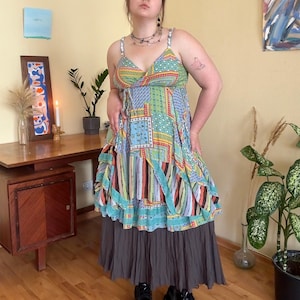 May include: A colorful patchwork dress with a V-neckline and spaghetti straps. The dress features a mix of patterns, including squares, stripes, and floral designs, in shades of blue, green, red, and orange. The skirt is a solid gray color.