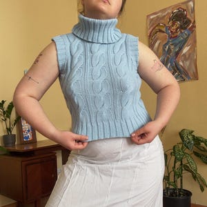 May include: A light blue cable knit sleeveless turtleneck sweater. The sweater has a ribbed hem and a high, folded collar. The knit pattern is a classic cable design. The sweater is worn with a white skirt.