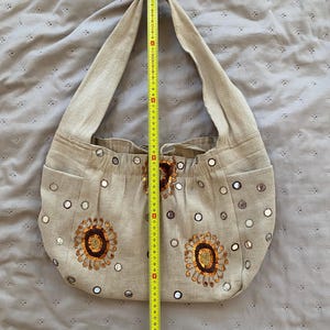 May include: A beige hobo bag featuring circular mirror embellishments and two embroidered floral motifs in brown and orange. A yellow measuring tape is placed vertically over the bag, showing a height of approximately 15 inches from top to bottom.
