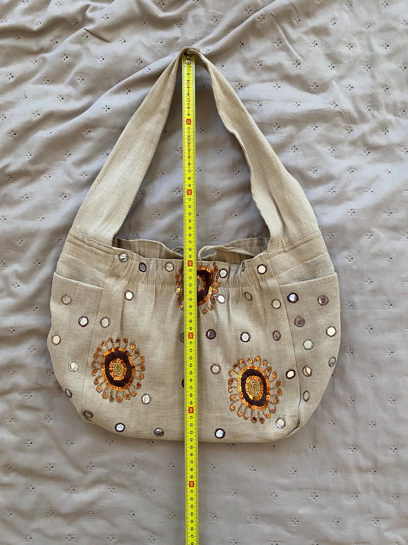 May include: A beige hobo bag featuring circular mirror embellishments and two embroidered floral motifs in brown and orange. A yellow measuring tape is placed vertically over the bag, showing a height of approximately 15 inches from top to bottom.