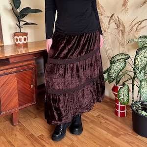 Puede incluir: Falda larga de terciopelo marrón oscuro con volantes y detalle de encaje. La falda se combina con una camiseta negra de manga larga y botas negras. La falda tiene un estilo bohemio.