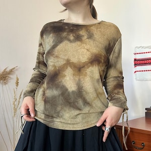 Puede incluir: Una blusa de manga larga con un patrón tie-dye en tonos marrón y bronceado con apariencia texturizada. La prenda tiene un cuello redondo y cordones laterales ajustables en los puños, creando un efecto fruncido en las mangas.