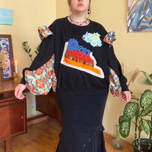 Könnte beinhalten: Schwarzes Langarm-Oberteil mit farbenfrohen Patchwork- und floralen Akzenten. Das Oberteil hat einen großen, dekorativen Aufnäher mit Schuhdesign und Spitzenbesatz. Die Ärmel haben ausgestellte, gemusterte Stoffdetails. Ein einzigartiges, künstlerisches Kleidungsstück.