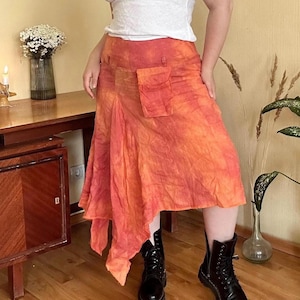 Może przedstawiać: Pomarańczowo-czerwona spódnica w stylu tie-dye z kieszenią. Spódnica ma asymetryczny dół i jest noszona z czarnymi butami. Spódnica jest wykonana z lekkiego materiału.