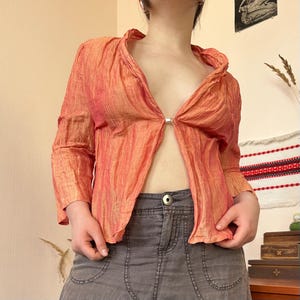 May include: A peach-colored, long-sleeved blouse with a textured, crinkled appearance. The blouse has a V-neck design and is secured with a silver-colored clasp. The sleeves have a slight ruffle at the wrist. The blouse is paired with a gray denim skirt.