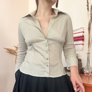 May include: A long-sleeved button-down shirt in a light beige color with thin vertical dark stripes. The shirt features a pointed collar, a fitted bodice with vertical seams, and a row of small buttons running down the center front.