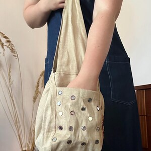 May include: A beige hobo-style shoulder bag featuring a single strap and an exterior pocket. The bag is decorated with numerous small, circular, reflective mirror-like embellishments arranged in a grid pattern across the front surface.