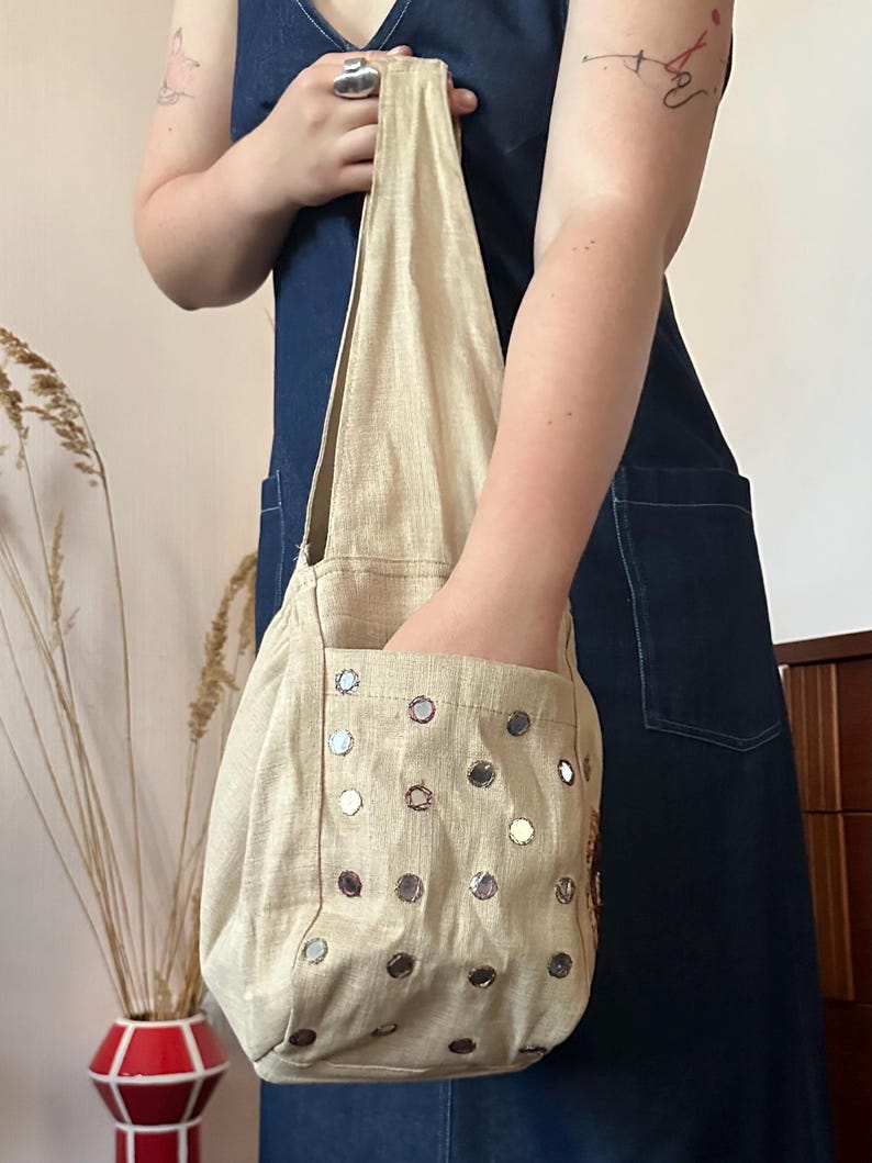 May include: A beige hobo-style shoulder bag featuring a single strap and an exterior pocket. The bag is decorated with numerous small, circular, reflective mirror-like embellishments arranged in a grid pattern across the front surface.
