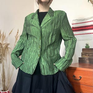 May include: A long-sleeved blazer in a vibrant green hue with a distinct vertical crinkled texture. The garment features a notched lapel collar, three matching green buttons down the front closure, and two front pockets, worn over a black top.