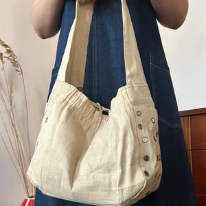 May include: A beige hobo-style shoulder bag made of a textured fabric. The bag features a gathered top opening with a drawstring closure and a wide shoulder strap. The sides are decorated with a vertical row of small, circular metallic mirrors.