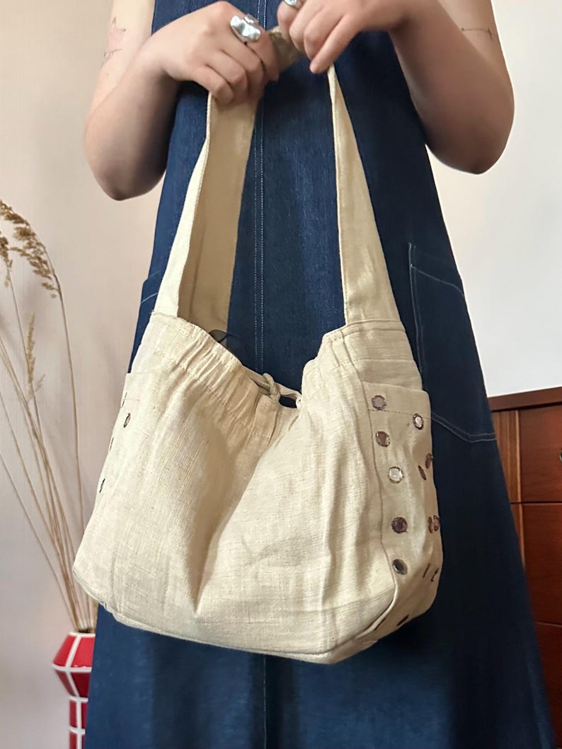 May include: A beige hobo-style shoulder bag made of a textured fabric. The bag features a gathered top opening with a drawstring closure and a wide shoulder strap. The sides are decorated with a vertical row of small, circular metallic mirrors.