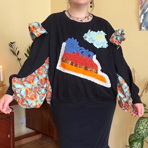 May include: A black long-sleeve sweatshirt featuring colorful patterned fabric inserts on the sleeves and shoulders. The front is decorated with a large, multi-layered appliqué made of blue and red lace, orange fabric, and a light blue floral lace patch.