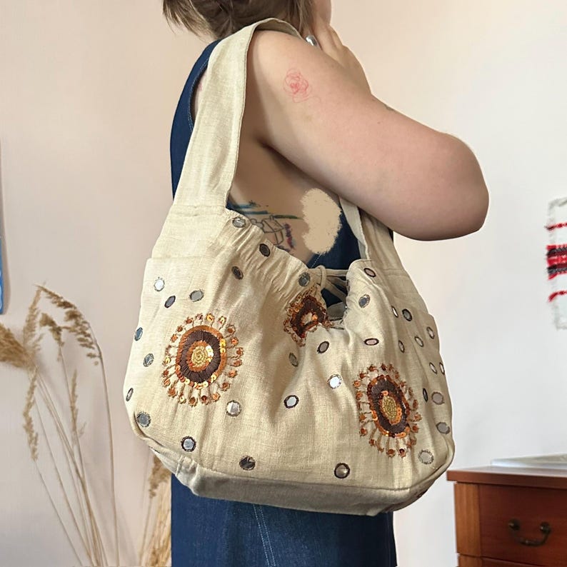 May include: A beige hobo bag featuring circular embroidered floral motifs in shades of brown and orange, accented with small reflective circular mirrors scattered across the fabric surface. The bag has a wide shoulder strap and a gathered top opening.
