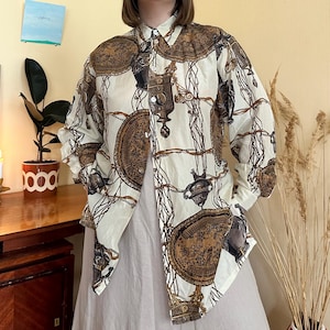 Może przedstawiać: Kremowa koszula zapinana na guziki z powtarzającym się wzorem brązowych medalionów, łańcuchów i elementów dekoracyjnych. Koszula z długim rękawem ma klasyczny kołnierzyk i guziki z przodu. Koszula jest rozpięta, odsłaniając neutralny kolor ubrania pod spodem.