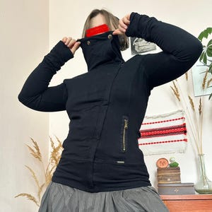 May include: Black jacket with a high collar and asymmetrical zipper. The jacket has long sleeves and a fitted silhouette. Gold-colored zipper and button details add contrast to the solid black fabric. The jacket is part of a clothing collection.