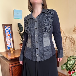 Può includere: Una camicia blu scuro con un design patchwork. Presenta un colletto, maniche lunghe e un mix di trame, inclusi pannelli a coste e fantasia. La parte anteriore ha diversi bottoni e toppe grigie.