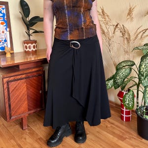 May include: A black midi skirt with a draped front and a silver-tone buckle. The skirt is paired with a patterned top in shades of brown and blue, and black boots. The outfit is styled in a vintage aesthetic.