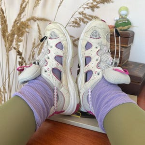 May include: Pair of off-white and grey athletic shoes with pink accents and mesh detailing. The shoes have a unique design with a lace-up closure and are worn with lavender socks. The shoes are on a wooden surface.