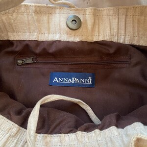 May include: A beige fabric bag featuring a dark brown interior lining, a zippered pocket, and a blue rectangular label with the white text ANNAPANNI. The bag includes a magnetic snap closure and a drawstring cord for securing the opening.