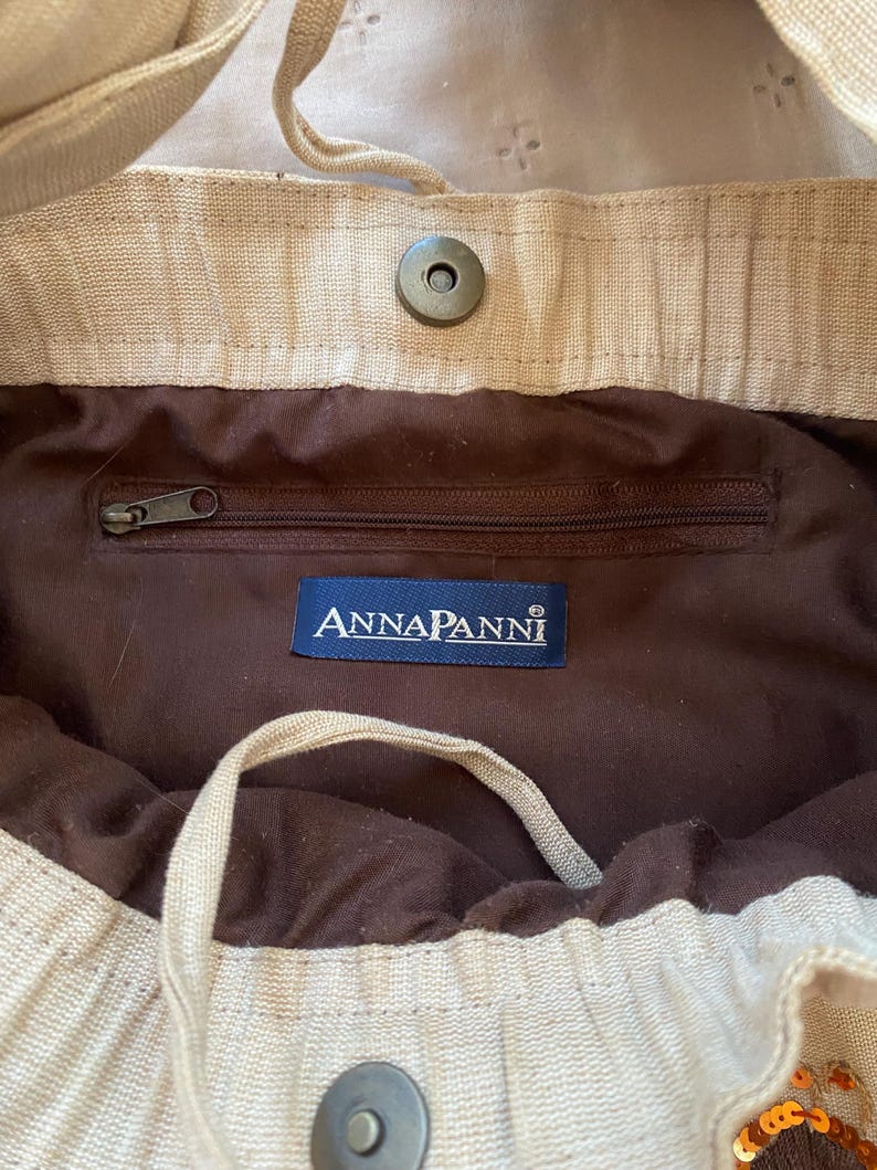 May include: A beige fabric bag featuring a dark brown interior lining, a zippered pocket, and a blue rectangular label with the white text ANNAPANNI. The bag includes a magnetic snap closure and a drawstring cord for securing the opening.