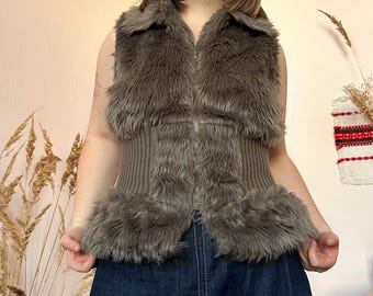 May include: A sleeveless, brown faux fur vest with a ribbed knit center panel and a zipper closure. The vest has a collared neckline and fur trim along the edges. The garment is worn over a dark blue denim skirt.