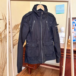 May include: Black jacket with a high collar, multiple zipped pockets, and buttoned flap pockets. The jacket has a vertical striped pattern and a hood. It is hanging on a wooden hanger.