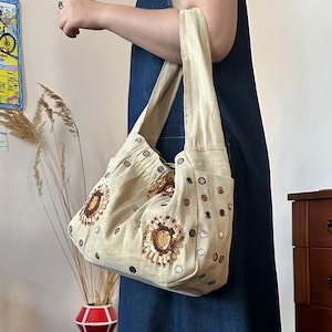 May include: A beige hobo-style shoulder bag made of textured fabric, featuring embroidered sunflower motifs with brown and gold sequins. The bag is decorated with small circular mirrors scattered across the surface and has a wide, integrated shoulder strap.