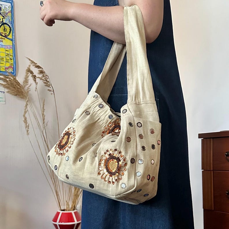 May include: A beige hobo-style shoulder bag made of textured fabric, featuring embroidered sunflower motifs with brown and gold sequins. The bag is decorated with small circular mirrors scattered across the surface and has a wide, integrated shoulder strap.